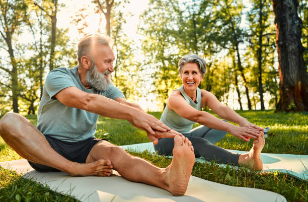 istockphoto-1912106674-612x612 Flexible exercises for body. Sporty man and woman with grey hair stretching on yoga mats with hands to one leg during outdoors workout. Happy married couple with bare feet warming up together at park.